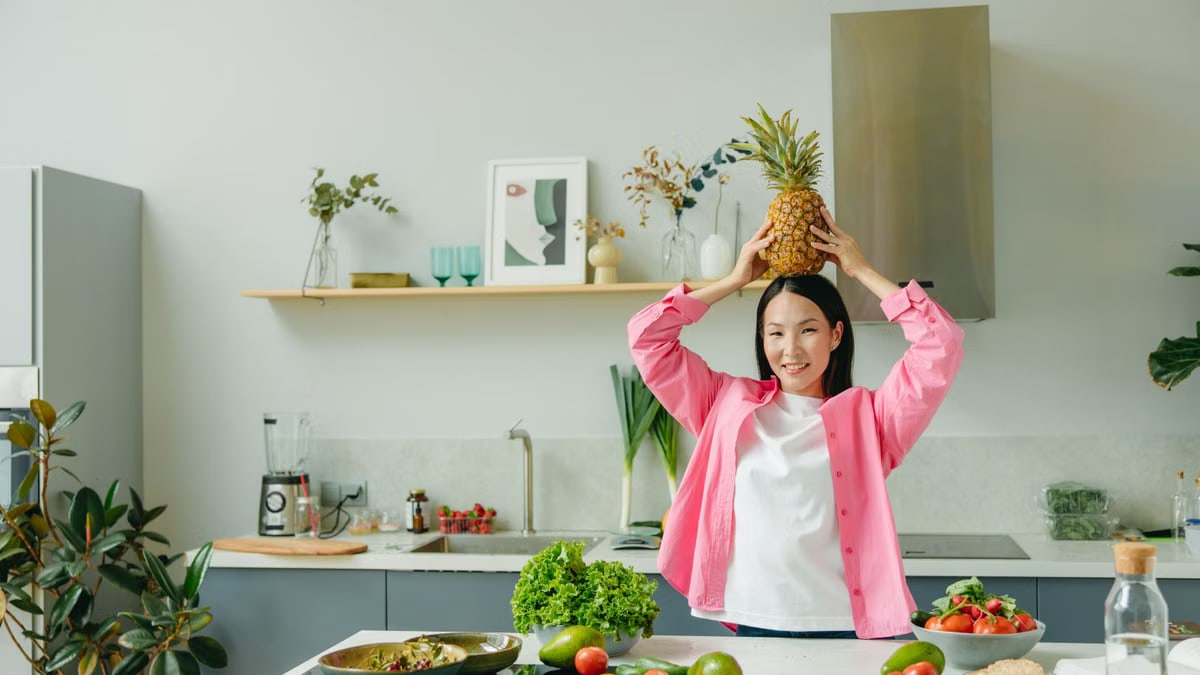 Asian woman in pink overshirt holding a pineapple in a modern kitchen with fresh produce and cheerful ambiance.